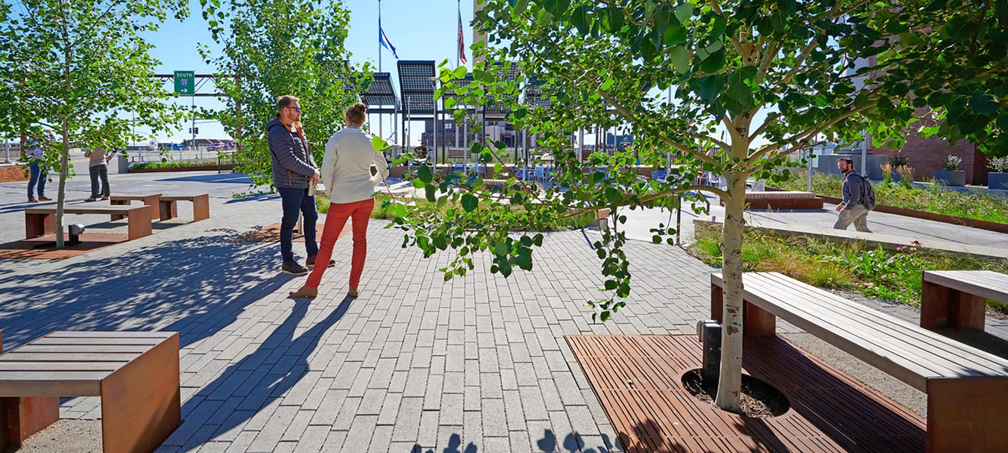 Two people standing amid benches and trees on a plaza made of pavers on a sunny day