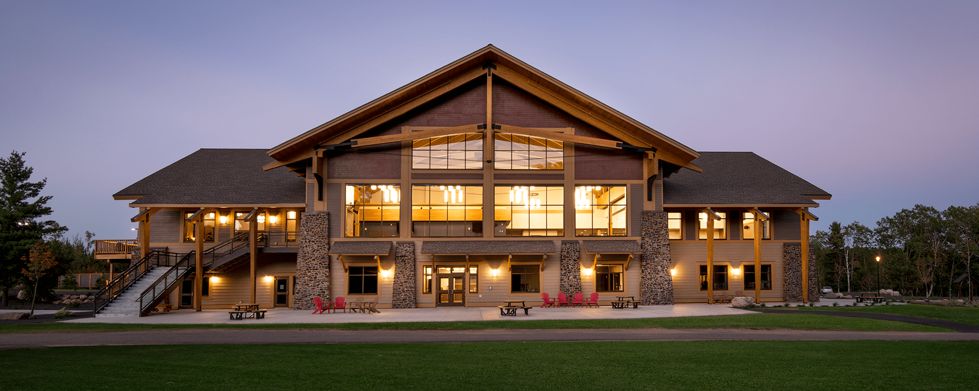 exterior view of lodge building with lights on inside. red chairs on green grass