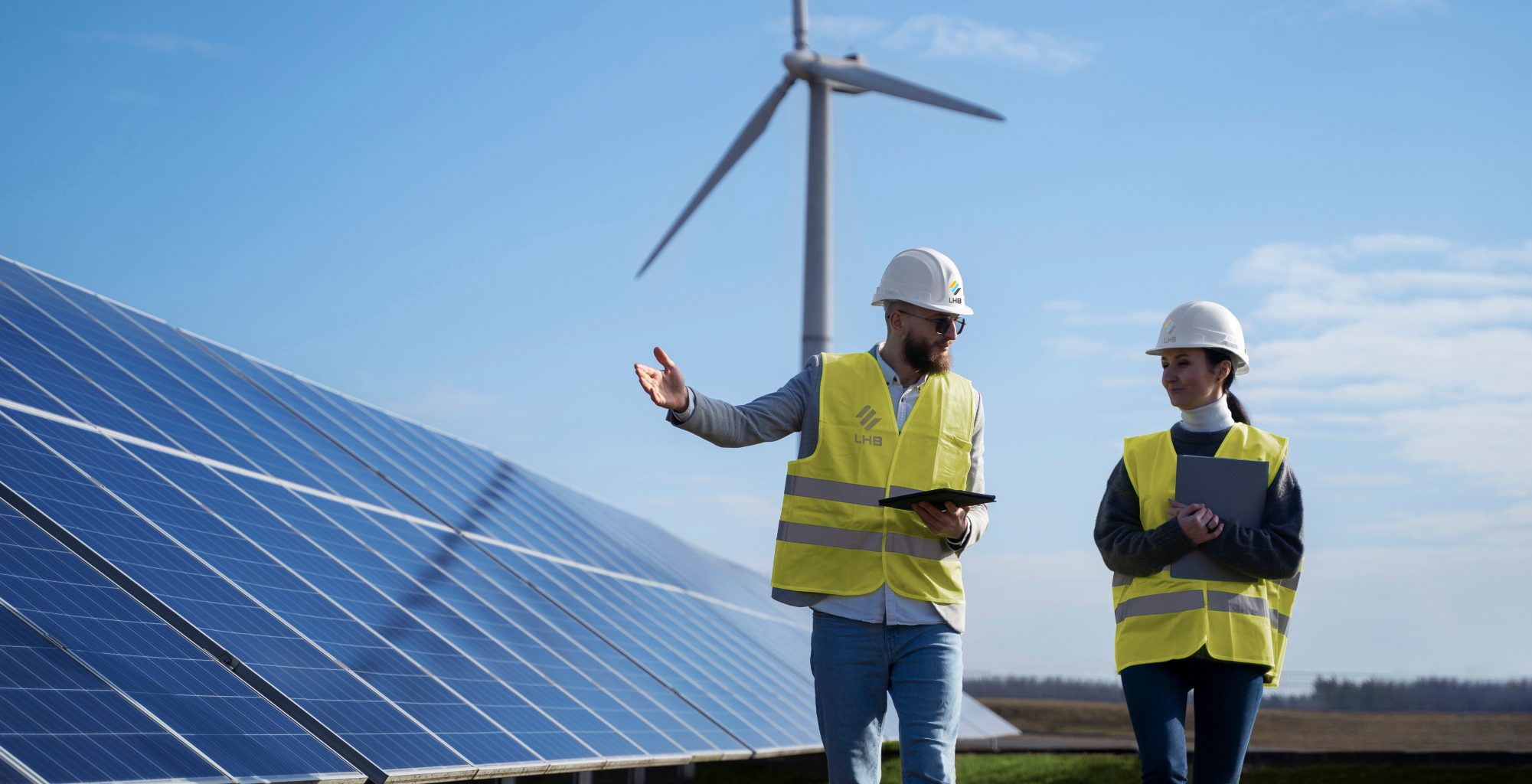 Workers in yellow vests near a solar panel under a wind turbine on a sunny day
